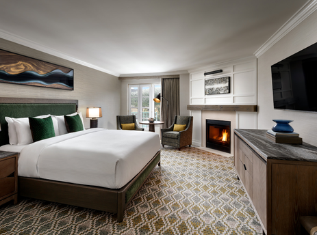 A luxurious bedroom featuring a king-sized bed with green accents, a fireplace, and a sitting area by a window with an exterior view.