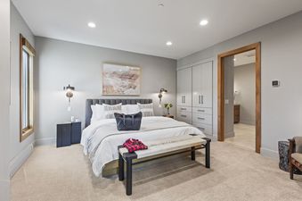 The primary bedroom features a comfortable king-size bed with crisp white linens and decorative pillows. The room is decorated in neutral tones with light grey walls, light-colored carpeting, and wooden accents around the window and doorway. There are bui