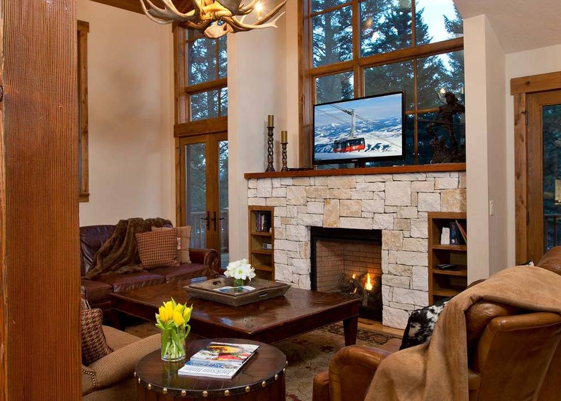 Cozy living room with stone fireplace, leather armchairs, and tall windows looking out to pine trees.