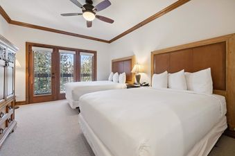 A hotel bedroom at The Charter C1205 is shown with two queen beds featuring white bedding and brown leather headboards. A ceiling fan hangs overhead, and double doors with stained glass lead to an outdoor view.