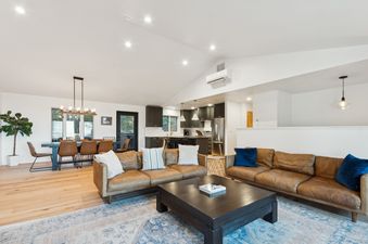 An expansive great room in Stumptown House features an open-concept layout, seamlessly connecting the living area with two brown leather sofas and a dark coffee table, the dining area with a dark wood table and tan chairs, and a modern kitchen with dark c