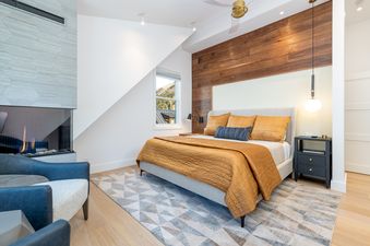 A bedroom with a fireplace, a bed with a gold comforter, a large window, and a ceiling fan.