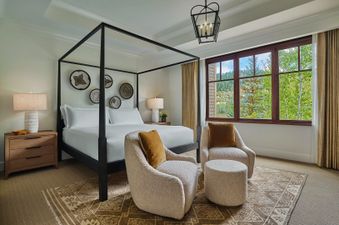 The primary bedroom features a striking four-poster king-sized bed, a pair of inviting armchairs, and a large window offering a view of the outdoors.