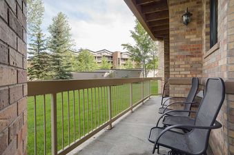 A private balcony at Chateau Chamonix CX313 with two black chairs, a beige railing, and a view of a green lawn, trees, and distant buildings under a cloudy sky.