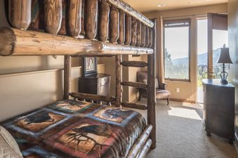 A rustic bunk bedroom with a log bunk bed, a patterned comforter, a leather armchair, a dresser with a lamp, and a large window offering mountain and forest views.