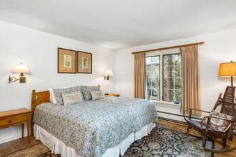 A cozy bedroom with a patterned duvet, floral throw pillows, two wall-mounted lamps, and a wooden rocking chair, all bathed in natural light from a large window.