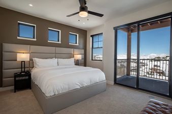 The Penthouse in the Pines features a king-sized bed with white linens and a padded headboard, accompanied by two nightstands with lamps and access to a balcony with mountain views.