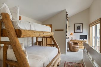 A rustic bunk room at Vail 21 406 features a wooden bunk bed with white bedding, a plush brown armchair, and a TV on a wooden dresser.