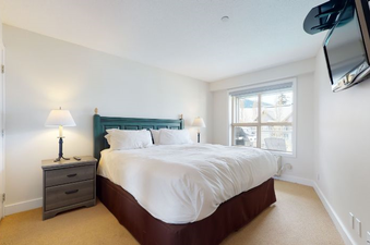 The comfortable bedroom in Aspens on Blackcomb Unit 461 features a large bed with white linens and a wooden headboard, two bedside tables with lamps, and a window offering a view of the surrounding area.