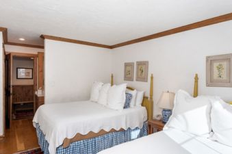 A bright bedroom in Chateau Christian 2 Bedroom Gold Unit 110 features two beds with white linens and blue patterned bedskirts, with a wooden nightstand and lamp between them.