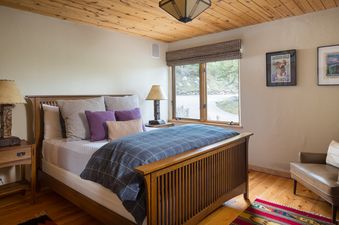 A cozy bedroom with a wooden bed frame, colorful pillows, and a large window overlooking a natural landscape.