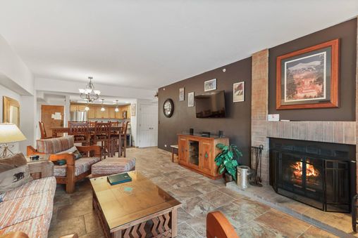 A living room with a fireplace, a large couch, two chairs, a coffee table, a large TV, and a dining area in the background.