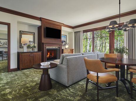 A luxurious living area with a fireplace, comfortable seating, and a dining table, featuring a large window with a view of lush greenery.