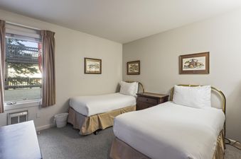 A cozy bedroom at Snow Flower 135 Park City featuring two twin beds with white linens and brass headboards, a nightstand, framed artwork, and a window with an air conditioning unit.