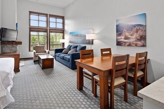 A spacious and inviting living area at Blackcomb Springs Studio features a blue sofa, a dining table, and a fireplace, with large windows offering a view of lush greenery.