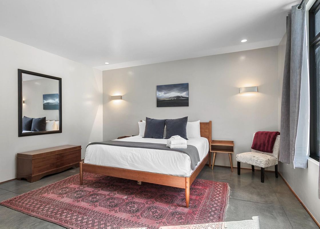 A serene bedroom features a wooden queen bed with white linens and dark grey pillows, flanked by modern wall sconces and a landscape painting, alongside a large mirror, a wooden chest, and an accent chair, all set on a concrete floor with a red patterned