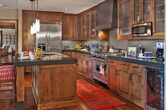 The image shows a large open plan kitchen area with a wooden kitchen island, wooden cupboards, and modern appliances. There is a large steel refrigerator on the left side of the kitchen island.