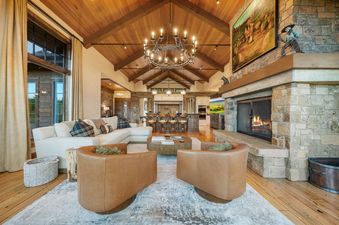 A luxurious and spacious living room with high wooden ceilings, a large stone fireplace, and elegant furnishings, including a white sectional and two brown leather armchairs.