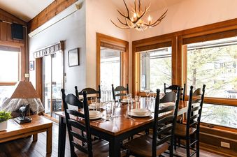The wooden dining table and chairs in Nez Perce A6 under a rustic antler chandelier next to large bright windows.