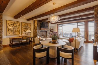 A luxurious living area at Peaks Edge at See Forever features a stone fireplace, plush white sofas, and a dining table, all illuminated by a unique chandelier and natural light from large windows offering an exterior view.