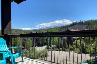 The outdoor balcony offers a breathtaking mountain view with lush green trees and distant snow-capped peaks under a blue sky.