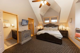 A master bedroom at Apres on French Street features a king-sized bed with a dark wood frame, soft white and blue striped bedding, and two nightstands with patterned lamps. A flat-screen television is mounted on the wall opposite the bed, and a partially v