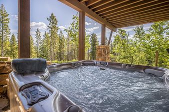 A close-up view of The Last Best Chalet's outdoor hot tub filled with bubbling water, with a control panel and a rolled towel visible, surrounded by a natural forest setting under a bright sky.
