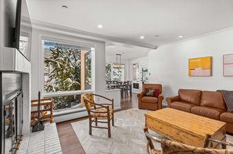 A living room with a leather couch, coffee table, and two chairs in front of a large window overlooking a snowy forest.