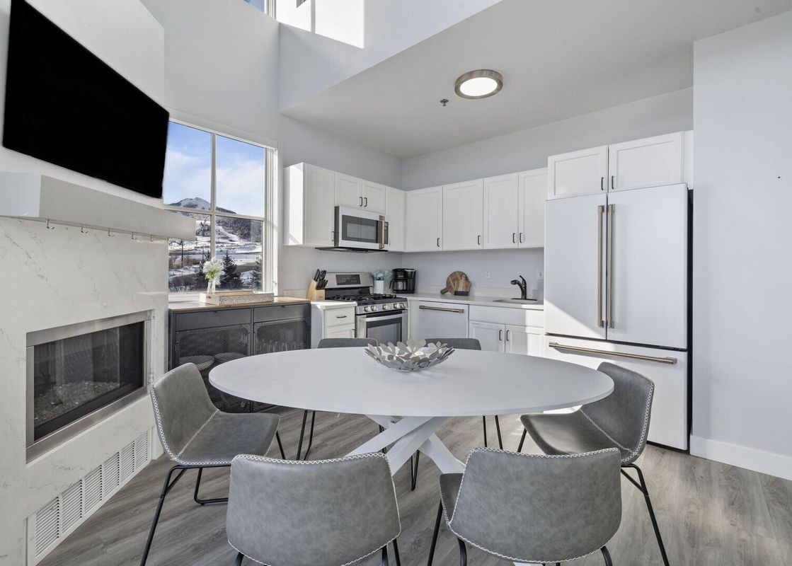 A modern kitchen and dining area with white cabinets, stainless steel appliances, a round dining table with gray chairs, and a window with a snowy mountain view.