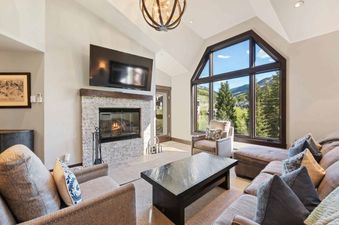 The spacious and elegantly furnished living room at The Charter B440 features a large fireplace, a flat-screen TV, and floor-to-ceiling windows offering stunning mountain views.