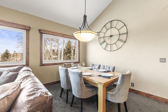 Dining table set for six with a large clock on the wall, a couch in the background, and windows offering a view of trees.