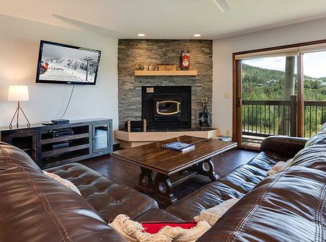 A cozy living room with a brown leather sectional, a fireplace, and a large window overlooking a scenic mountain landscape.
