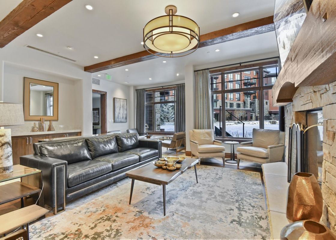 A spacious and inviting living area features a large black leather sofa, a rustic wooden coffee table with decorative bowls, and two light-colored armchairs, all set on a patterned area rug. Large windows offer a view of a snowy landscape and other buildi