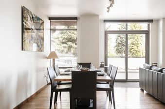 A modern dining area in Lowell 101 with a set table for six, bathed in natural light from large windows showcasing outdoor greenery.