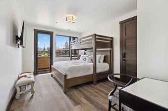 A modern bedroom with a wooden bunk bed, a flat-screen TV, a cowhide bench, a desk, and a balcony with an outdoor view.