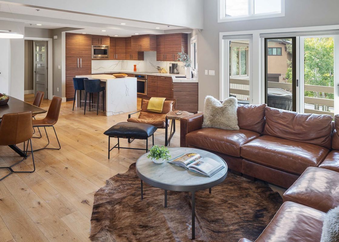 A spacious, open-concept living area with a brown leather sectional, a modern kitchen with a white marble island, and a dining space with a dark wood table and leather chairs, all brightly lit by natural light.