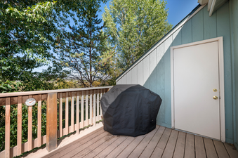 The outdoor living space at Twin Rivers Vasquez 3 features a grill covered in black, a wooden deck, and a privacy wall, all set against a lush green treeline.