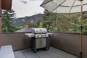 A private balcony with a Weber Genesis grill, shaded by a white umbrella, overlooking a mountain landscape.