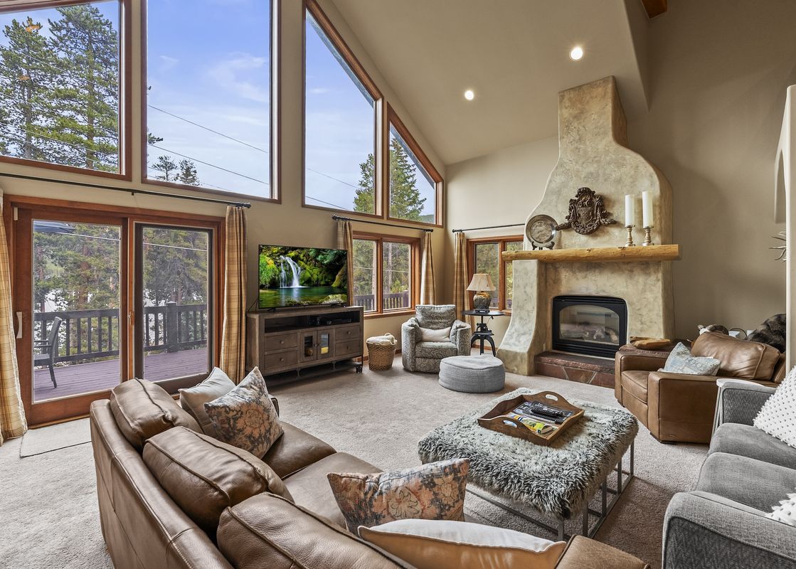 A spacious and luxurious living room with high ceilings, large windows offering a view of pine trees and a lake, a large brown sectional sofa, a stone fireplace, and a flat-screen TV.