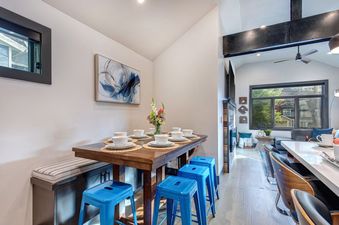 A dining room with a wooden table and blue stools, an open kitchen, and a living room with a fireplace.
