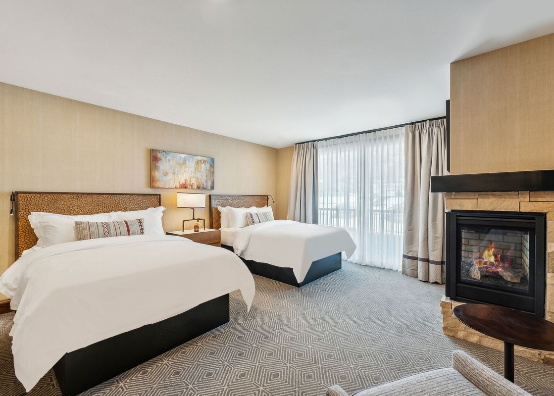 A luxurious bedroom featuring two queen-sized beds with white bedding, a decorative headboard, and a cozy fireplace, all set against neutral tones and elegant drapes.