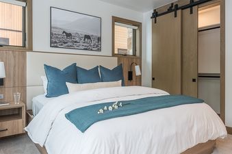 A bright bedroom at Riverlight Lodge features a large bed with white linens, blue pillows, and a teal throw, flanked by wooden nightstands and modern wall sconces, with a large horse artwork above the headboard and barn doors leading to a closet.