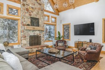 A cozy living room in Palmyra 94 Mountain Tranquility features a stone fireplace, comfortable seating, and large windows looking out onto a snowy landscape.