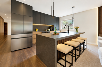 A modern kitchen with dark cabinetry, a large grey stone-topped island, and four beige barstools in Montaneros 302.