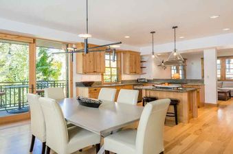 A dining area with a large table and chairs in front of a kitchen with wood cabinets and a large window overlooking a balcony.