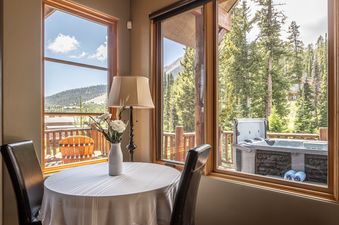 A cozy dining area at Moonlight Mountain 9 Happy Trails with a round table, chairs, and large windows looking out to a forest, mountains, and a hot tub on the deck.