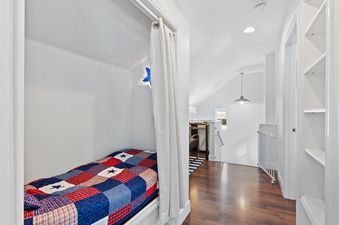 A cozy sleeping nook with a red, white, and blue quilt on a bed, next to a hallway with dark hardwood floors and an industrial pendant light.