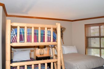 A cozy bedroom with a wooden bunk bed on the left and a single bed on the right, featuring colorful plaid bedding and a warm, inviting atmosphere.