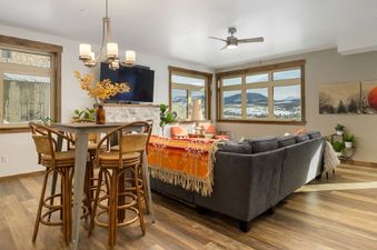 A living room in 448 Cranmer Unit 103 is shown with a gray sectional sofa, a small bar-height dining table with stools, and large windows revealing a snowy mountain landscape.