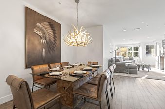 A modern dining room featuring a glass-top table with a wooden base, surrounded by brown leather chairs, under an antler-inspired chandelier.